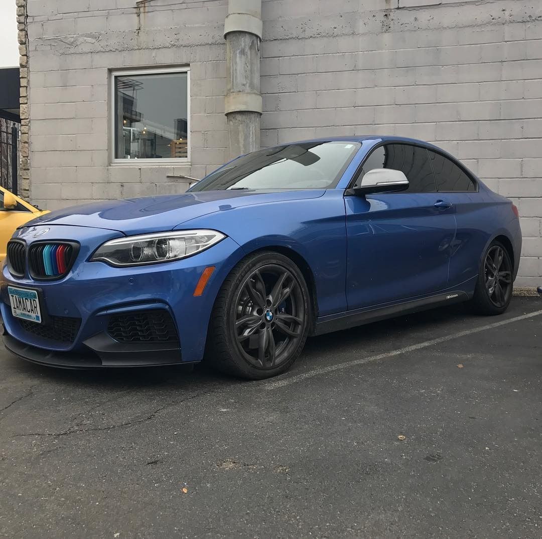 A blue BMW 2 Series coupe parked on asphalt in front of a grey concrete block wall.
