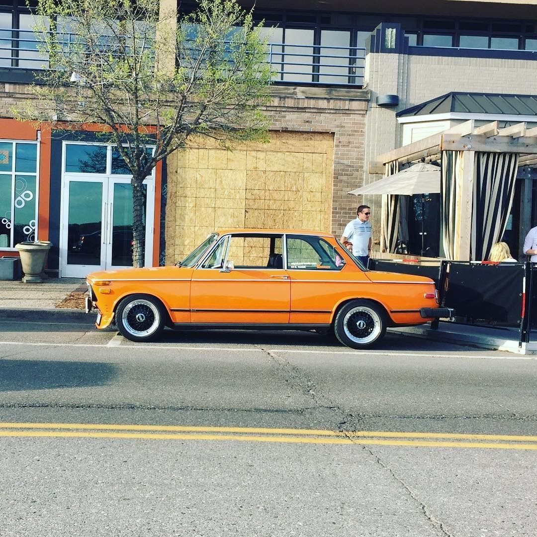 A bright orange vintage BMW coupe parked on the street in front of a brick building with a covered patio.