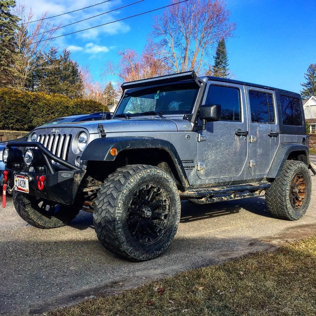 A grey four-door Jeep Wrangler with a lifted suspension, off-road tires, and a roof-mounted light bar parked outdoors.