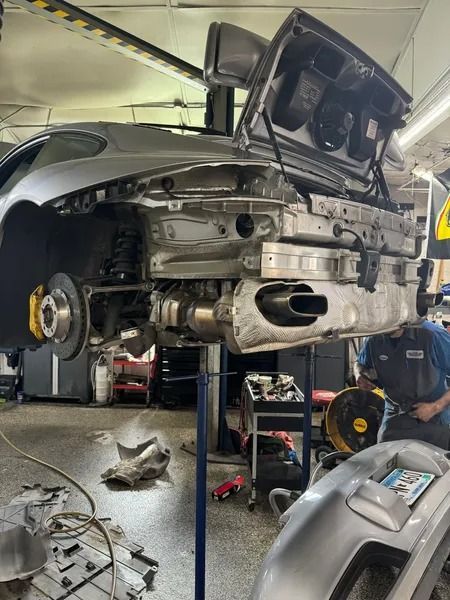 Silver car in a repair shop with its rear bumper removed, suspended on a lift while a mechanic works on the exhaust.