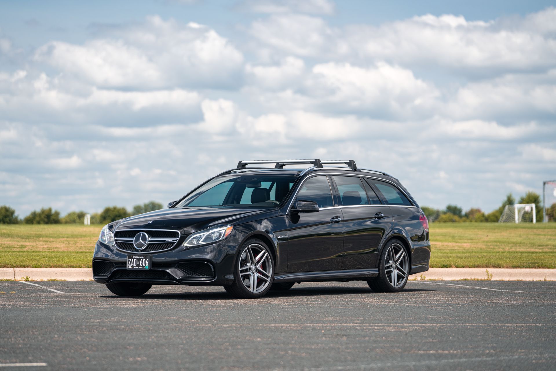 A black Mercedes-Benz station wagon with a roof rack parked on an asphalt lot under a cloudy blue sky.