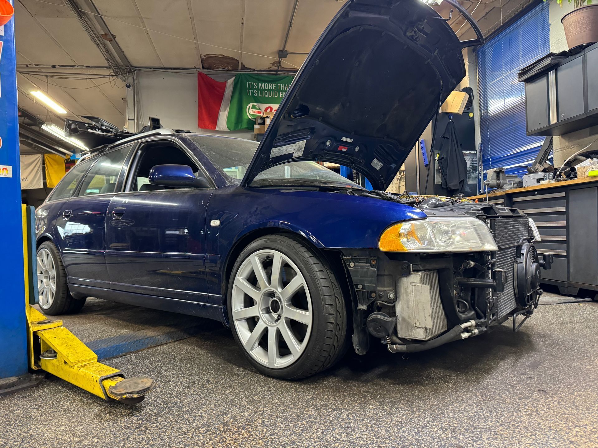 Blue sedan with hood open in an auto repair shop, front bumper removed, on a lift.