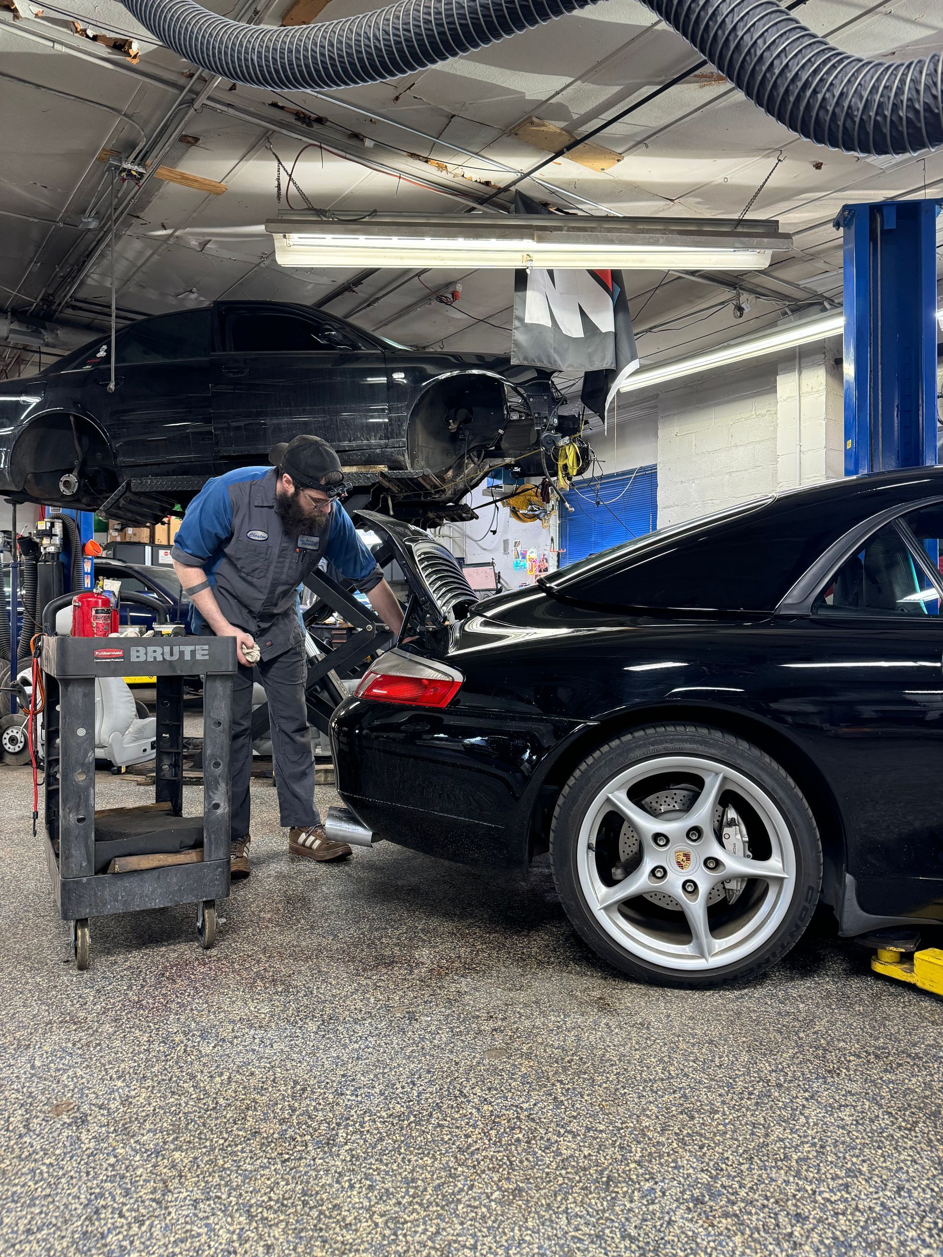 Mechanic working on a black sports car in a garage, with tools and equipment nearby.