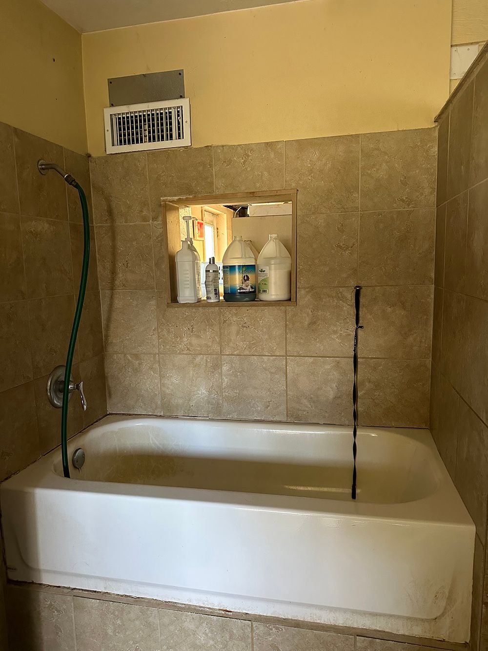 A tiled shower area with a white bathtub, a handheld shower hose, and a recessed wall niche containing bottles.