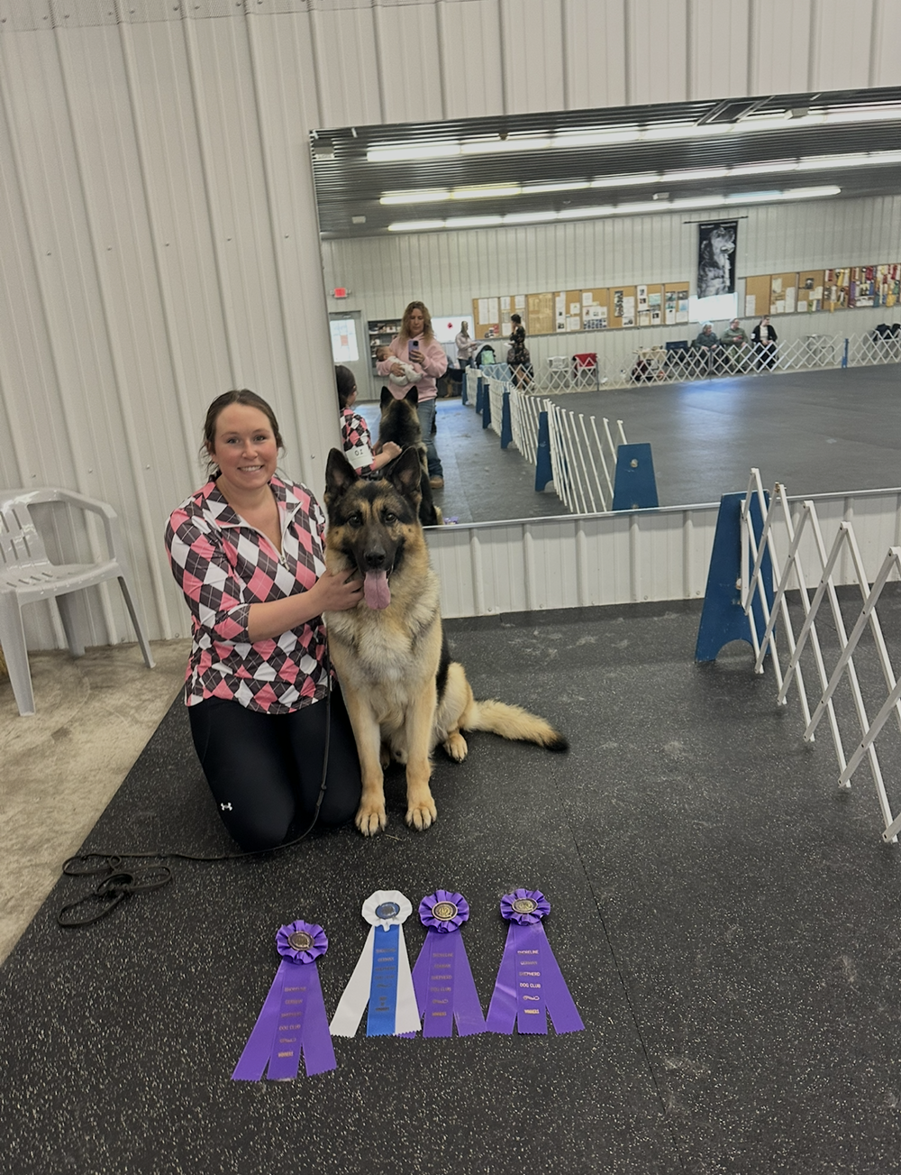 A person kneeling beside a German Shepherd in an indoor training facility, with four award ribbons displayed on the floor.