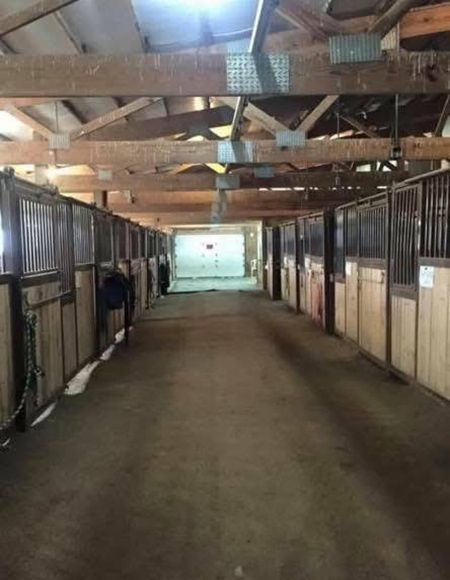 A wide aisle of an indoor horse barn with wooden stall fronts on both sides leading to a bright, open doorway.