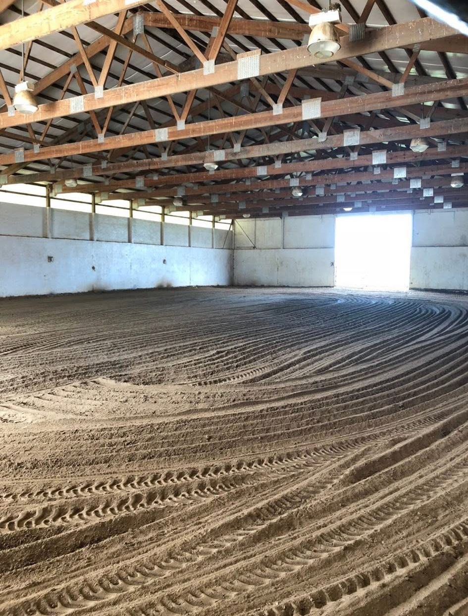An indoor equestrian riding arena with a sandy surface, exposed wooden rafters, and a bright open doorway at the back.