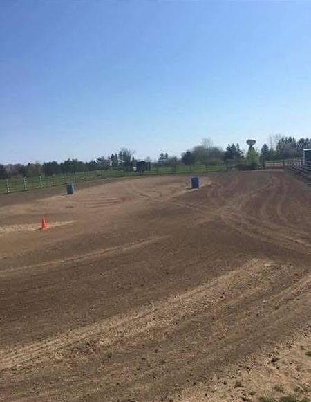 A flat, dirt training ring under a bright blue sky, featuring two blue barrels and one orange cone scattered in the field.