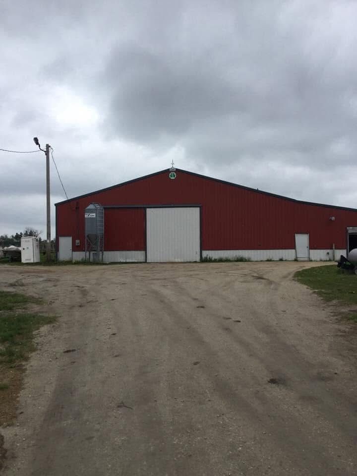 A red pole barn with a white sliding door, a side silo, and a dirt driveway under a cloudy sky.
