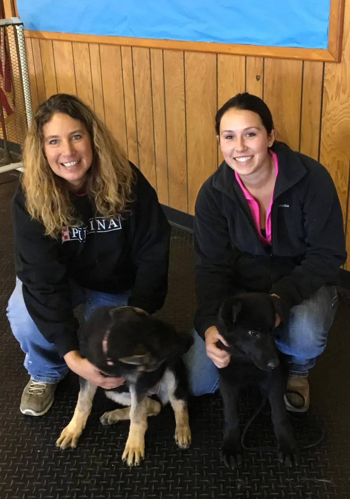 Two women kneeling with two dogs in front of wood-paneled wall, smiling.