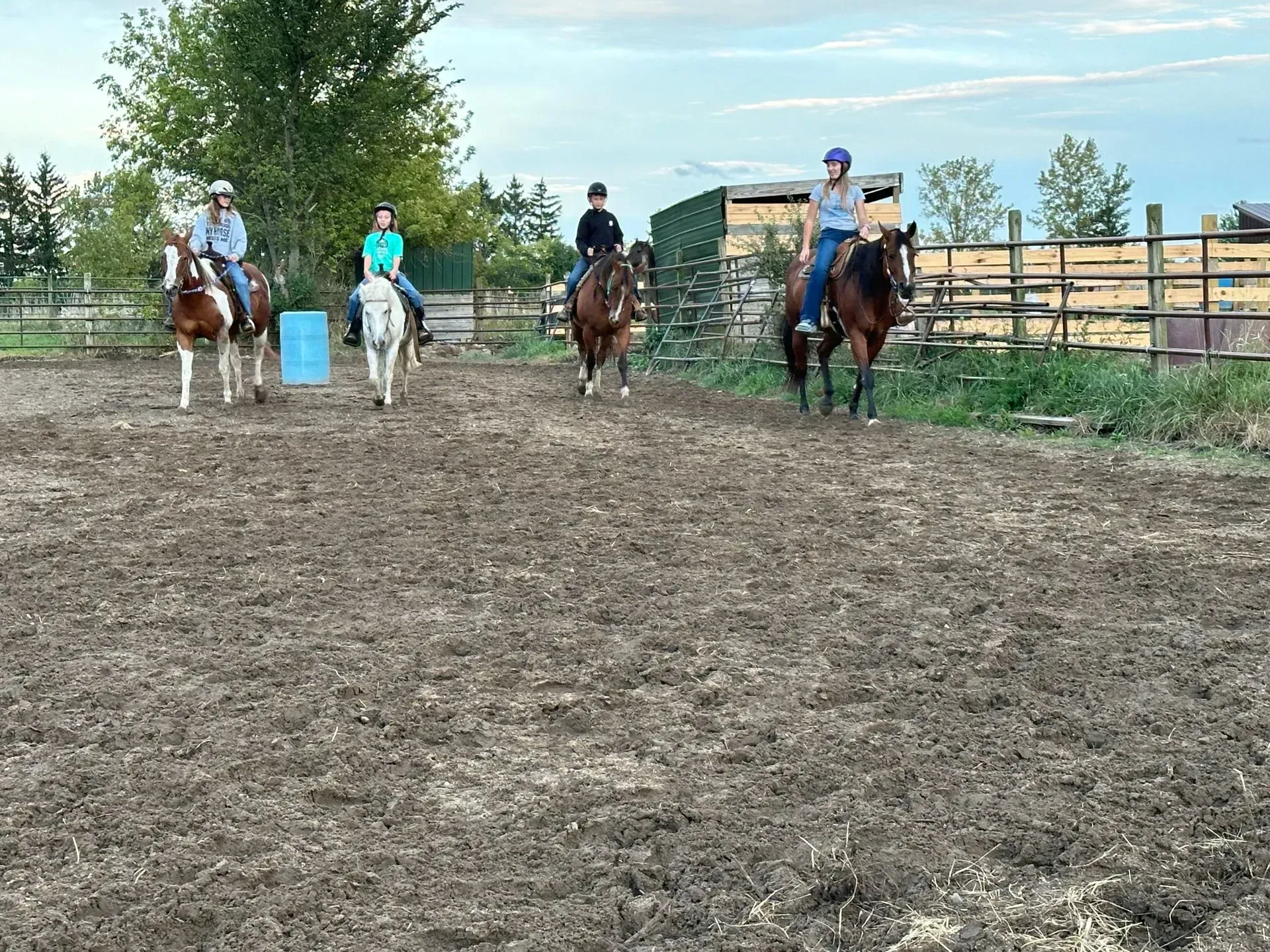 People riding horses in a muddy corral, near a wooden fence and trees, under a cloudy sky.