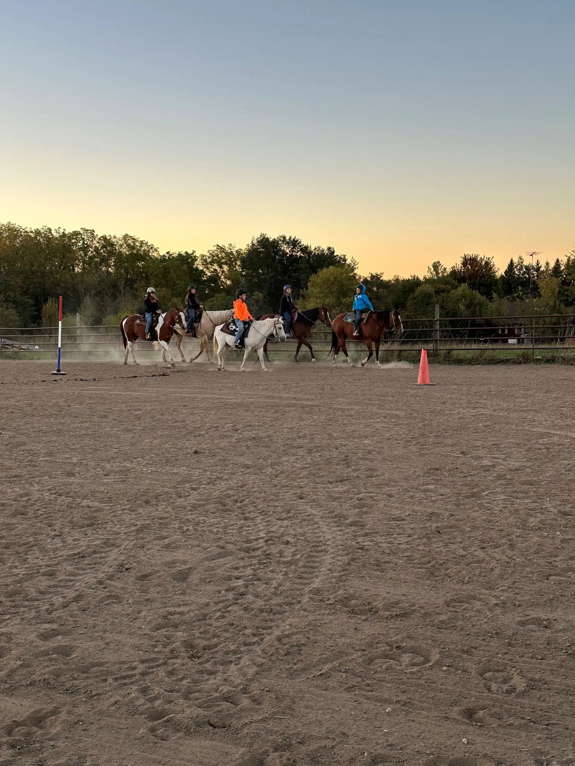 People riding horses in an outdoor dirt arena, with trees and a sunset in the background.