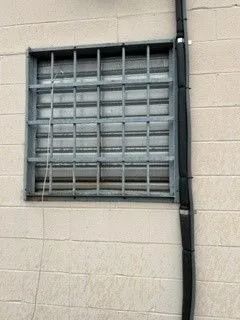Window with metal security bars on a beige brick wall, black rain gutter on the right.