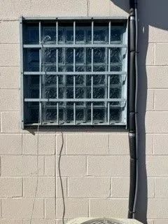 Window with security bars and glass blocks on a beige brick wall, next to a black pipe.