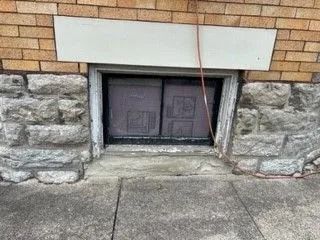 Basement window covered with cardboard, set in a stone and brick wall. Concrete sidewalk in front.
