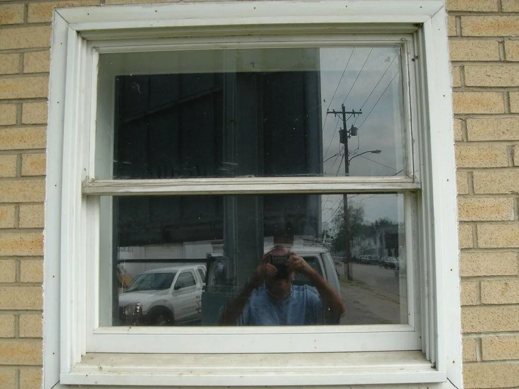 Window reflecting a street scene and person taking a photo; white frame and brick wall.