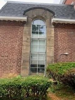 Tall arched window in brick building with gray roof and green bushes below.