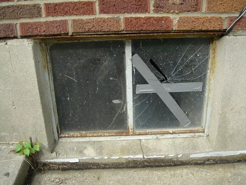 Broken basement window with duct tape, in brick wall.