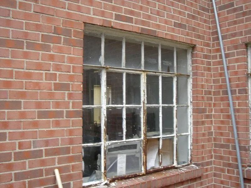 A weathered, multi-pane window in a red brick building; rusty white metal frame, some glass broken.