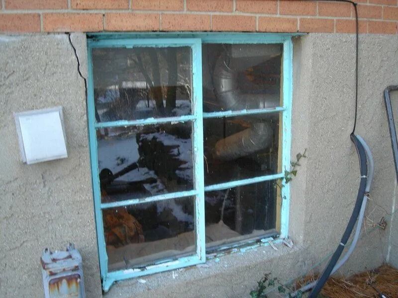 A window with peeling blue paint, set in a light stucco wall. A crack is visible at the top-left.