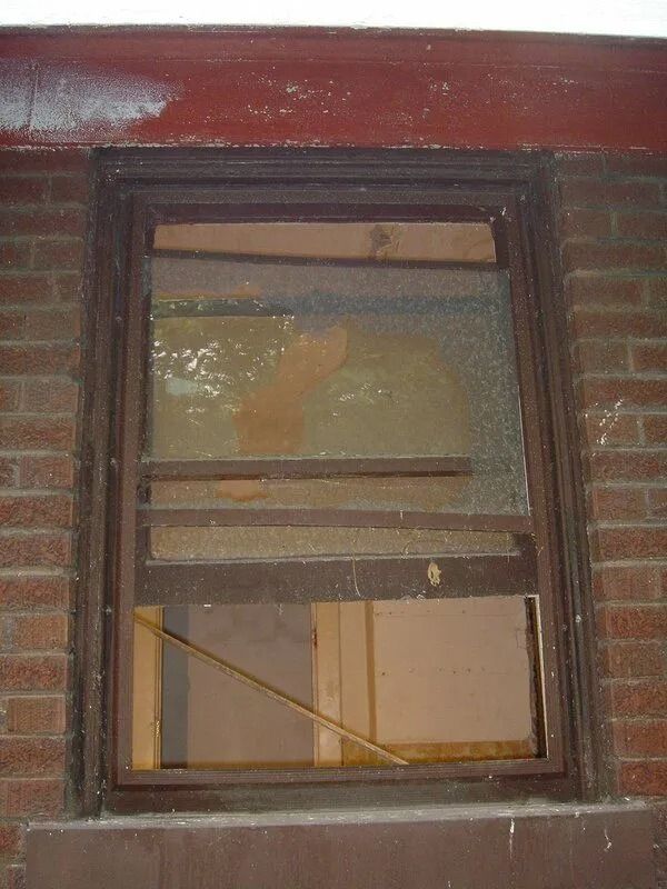 A boarded-up, broken window in a brick building. Wooden planks cover the glass; the frame is brown.