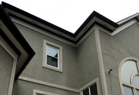 Exterior of a stucco building with a window. Black gutters and trim contrast the green-gray walls.