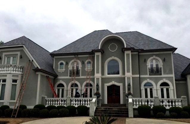Large, two-story house with gray exterior and dark roof; workers on ladders in front.