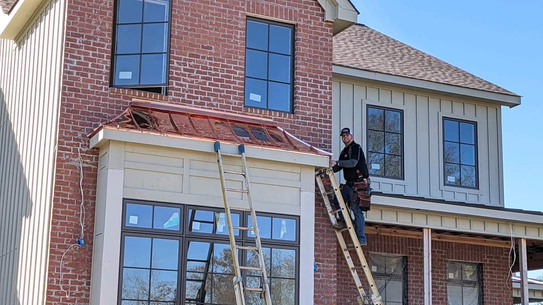 Man on ladder installing window on brick house with copper roofing.