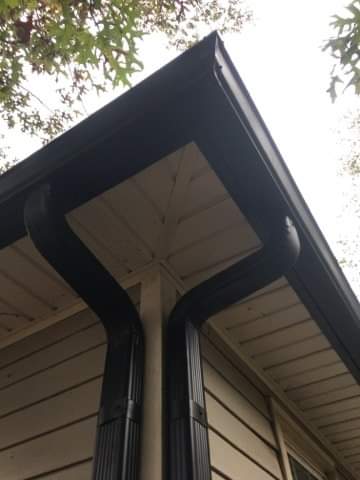 Black gutters on a house corner. Light-colored siding and a glimpse of foliage above.