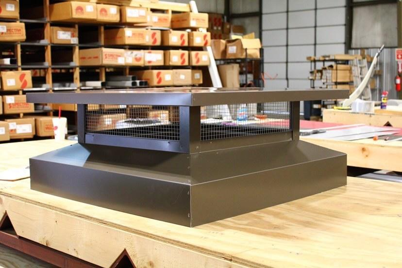 Chimney cap, brown metal, with wire mesh screen. On a wooden surface, industrial setting.