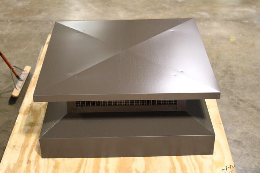 Square metal chimney cap, brown, with air vents, on a wooden base, sitting on a concrete floor.