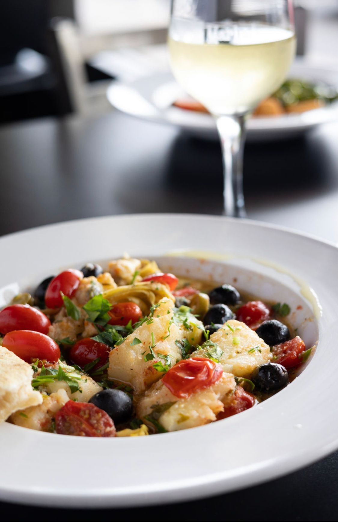 A bowl of food with tomatoes and olives next to a glass of wine.