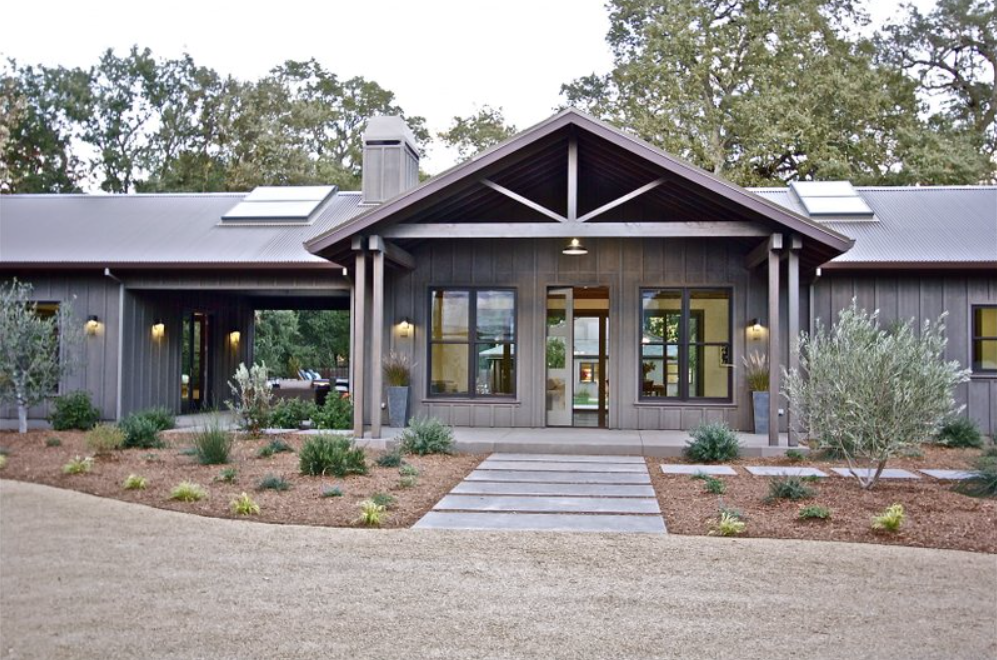 Gray-sided modern farmhouse with covered porch, wooden walkway, and olive trees in a gravel yard.