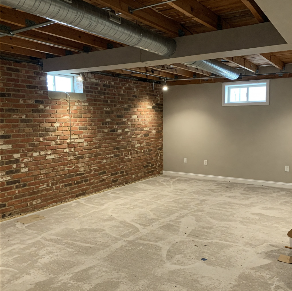 Interior of a basement with exposed brick wall, concrete floor, two small windows, and overhead ductwork.