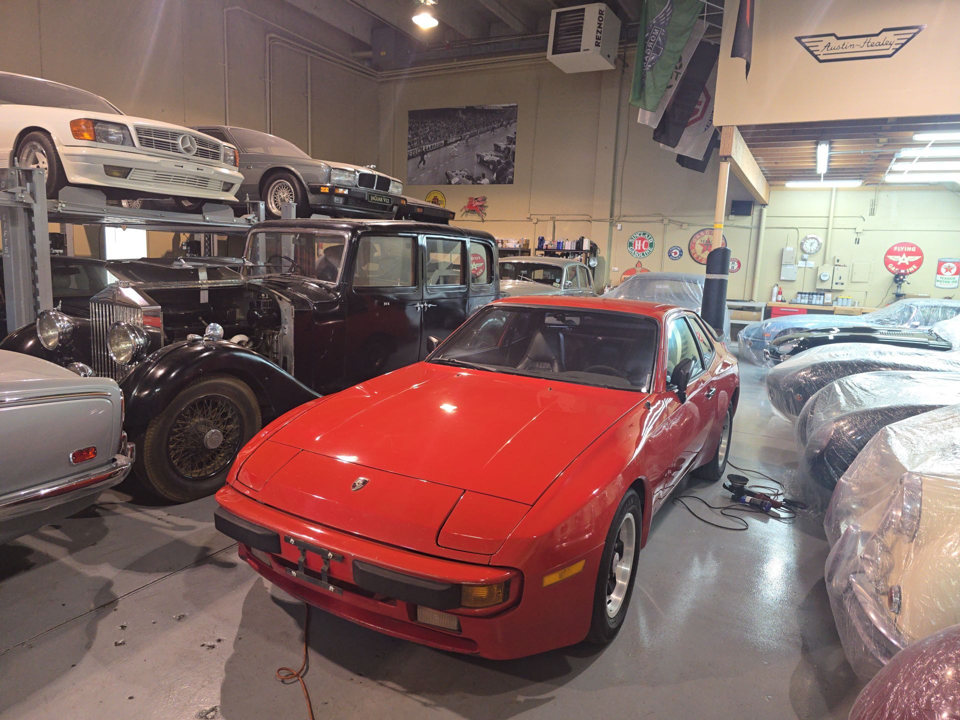 Red sports car in a cluttered auto garage with classic cars and parts around it