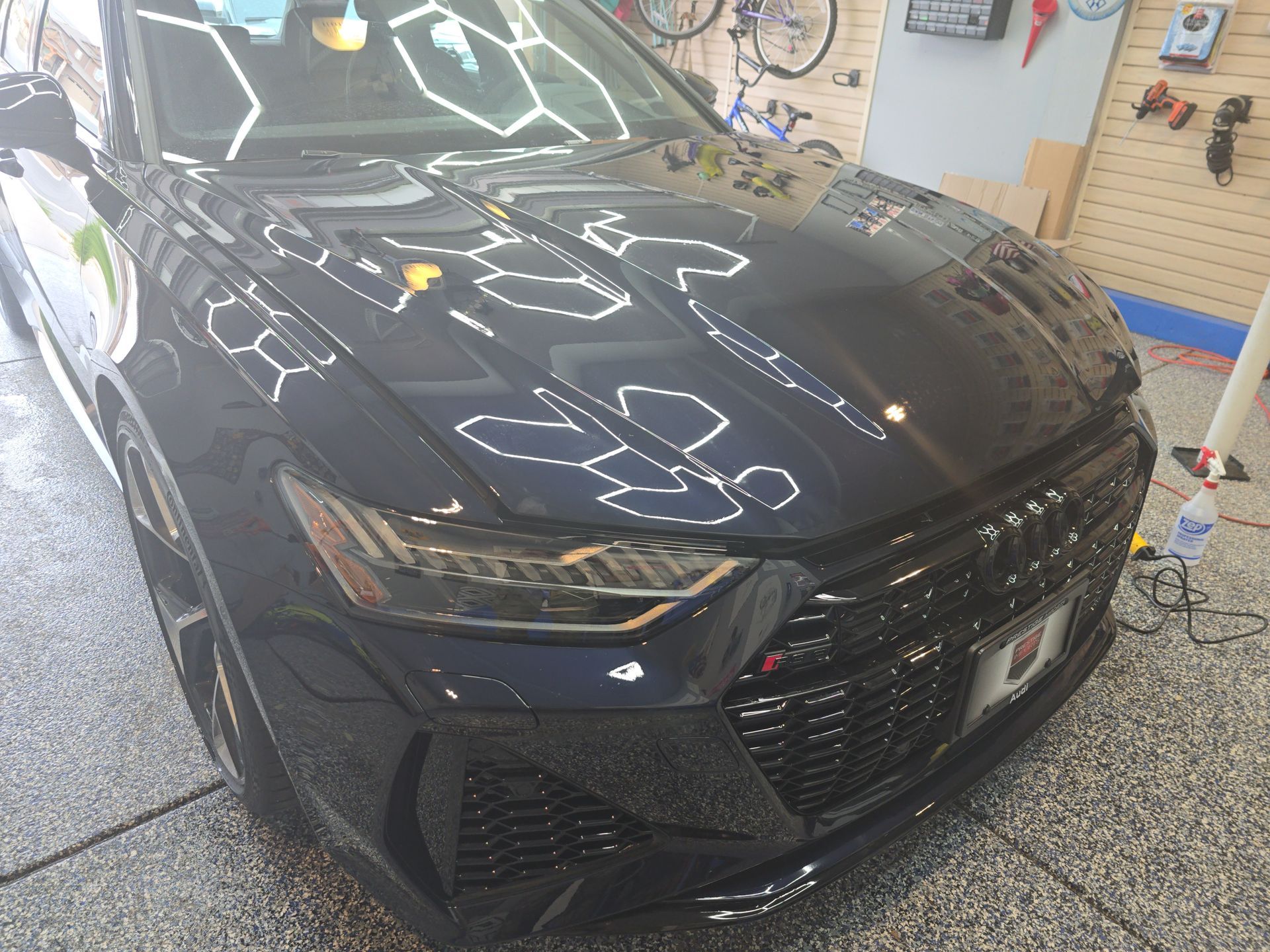 Black car in a workshop garage, front view with reflective hood and visible headlights.