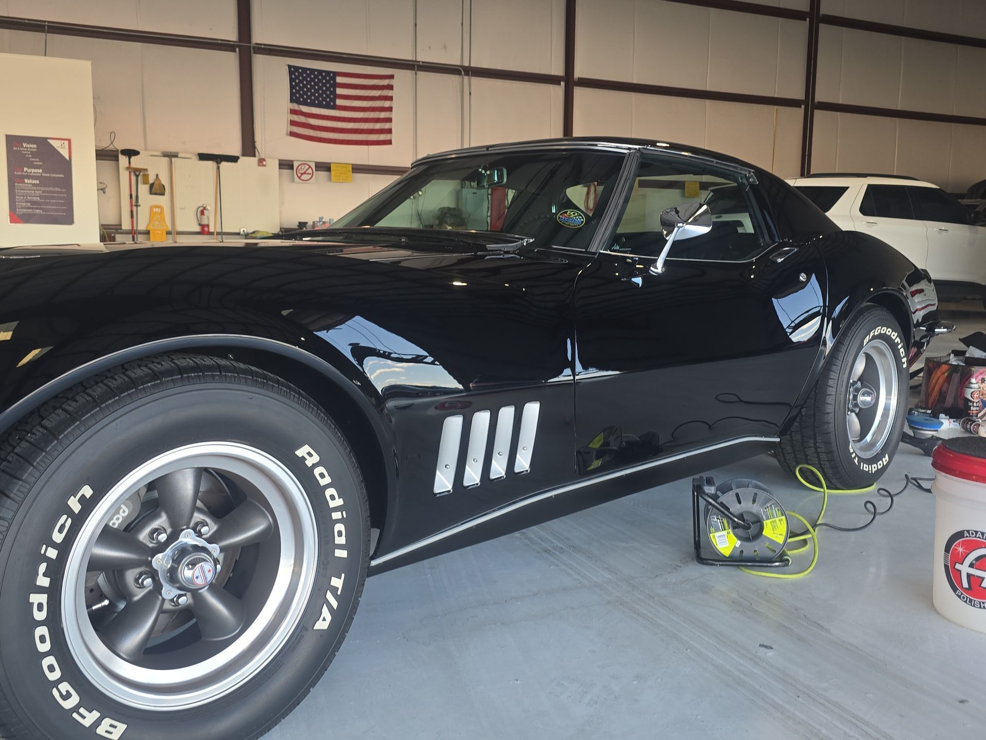 Black classic muscle car in a garage, with racing stripes and a charger on the floor nearby.