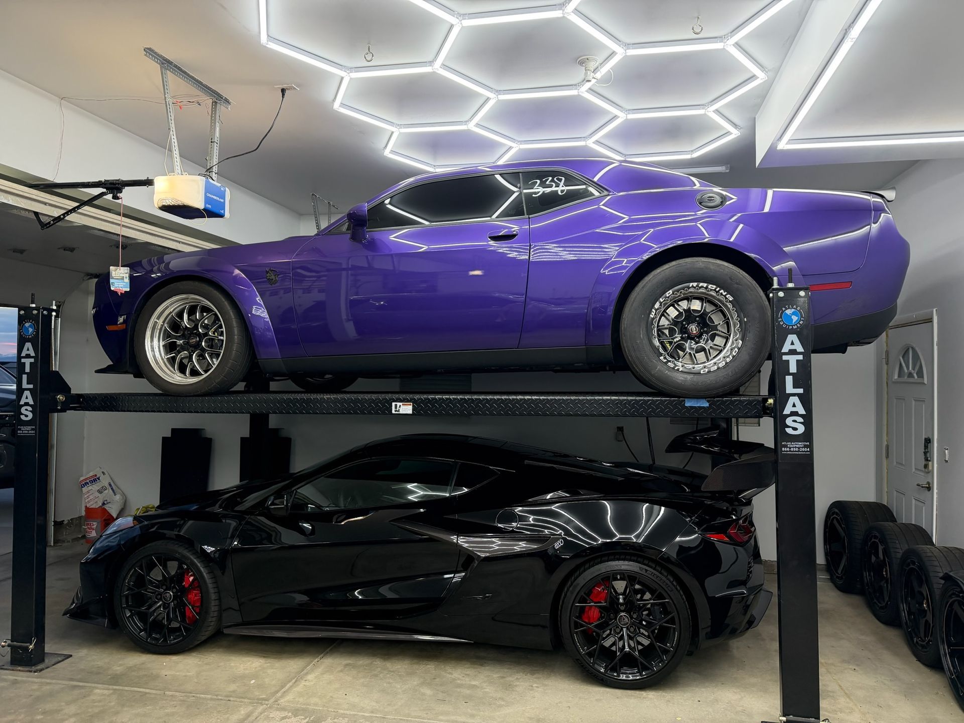 Two sports cars stacked in a garage: purple coupe on top, black coupe below, under geometric ceiling lights