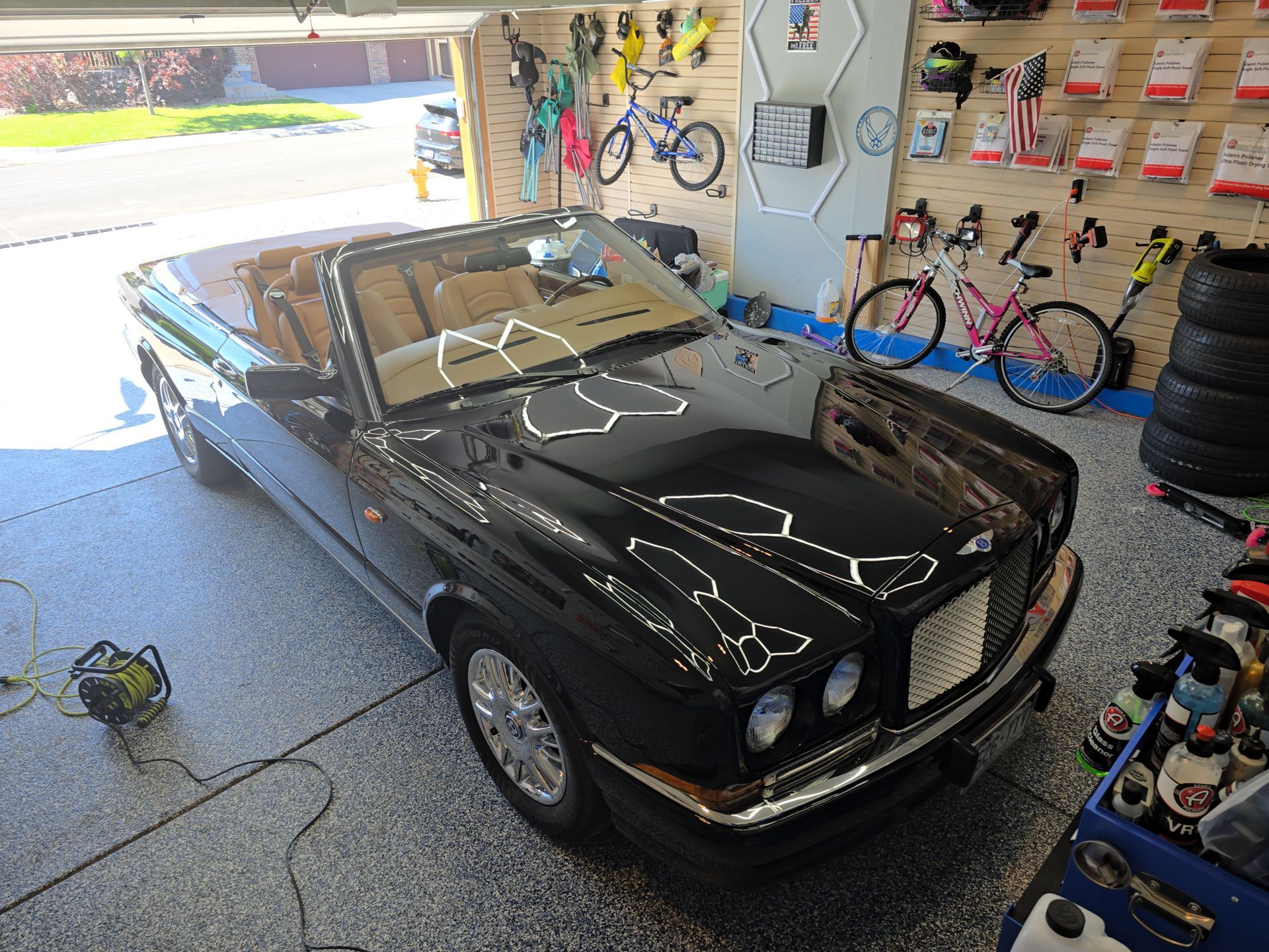 Black classic convertible parked in a garage, with bicycles and tools on the wall.