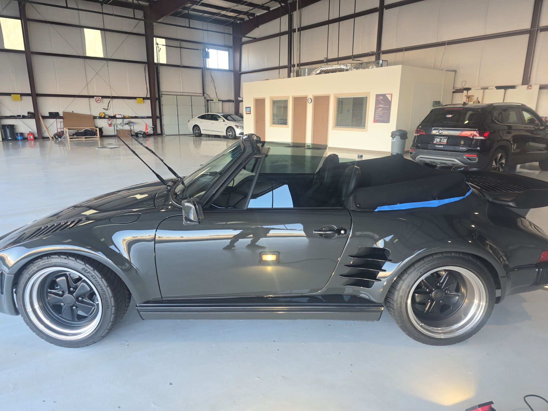Black convertible sports car parked inside a large garage with industrial walls and a black SUV in the background