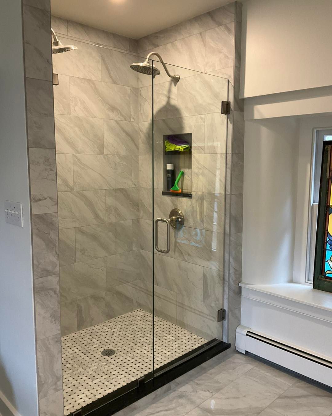 Modern shower with glass door and marble-look tile, inset shelf with toiletries, and small mosaic floor tiles.