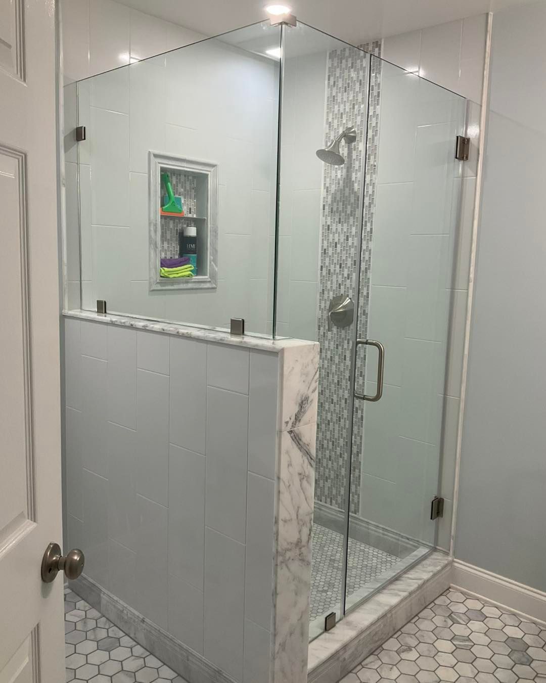 Glass-enclosed corner shower with a tile accent wall, set in a light blue bathroom with white tile flooring.