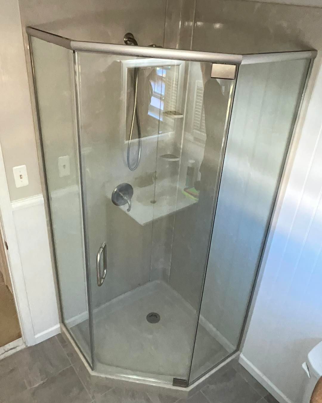 Corner shower with glass doors and silver frame. White and gray tile, built-in shelf.
