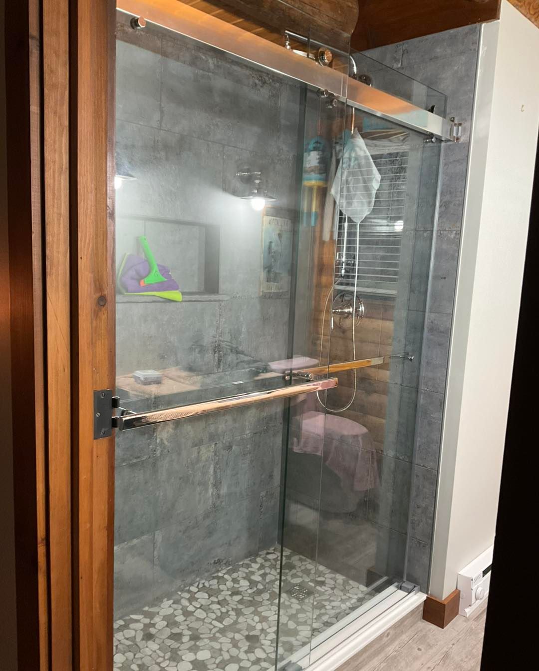 Shower with sliding glass doors, gray tiled walls, stone floor, wooden trim.