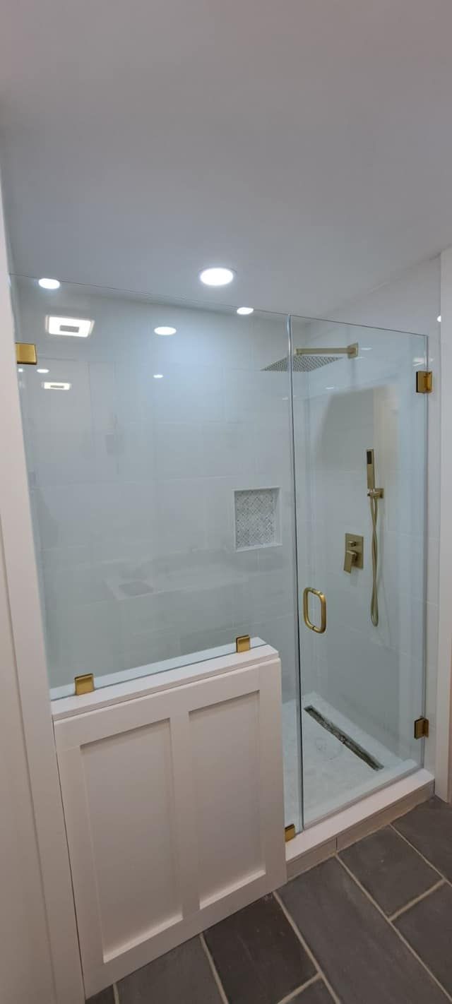 Modern bathroom with glass shower enclosure, white tile walls, and gold fixtures. White paneling in front.
