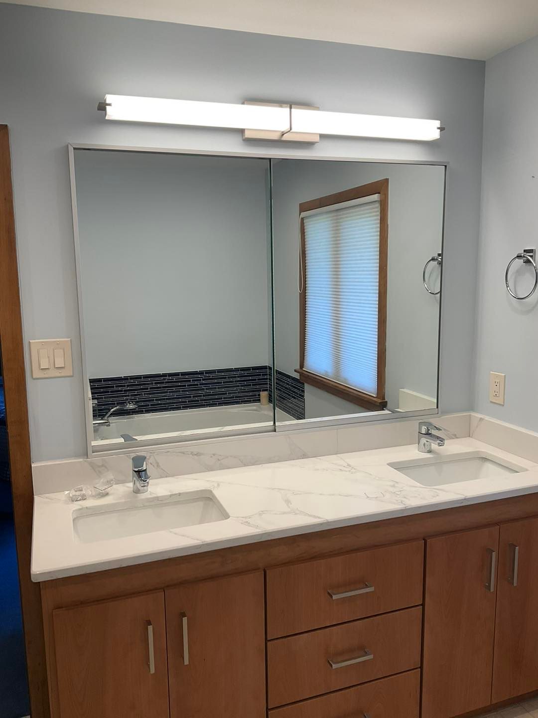 Bathroom with light blue walls, wood vanity with marble countertop, two sinks, large mirror, and window.
