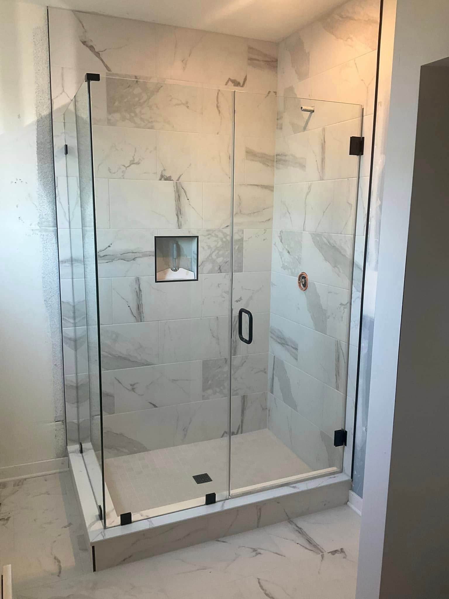Glass-enclosed shower with white marble-look tiles and black hardware, set in a light-colored bathroom.