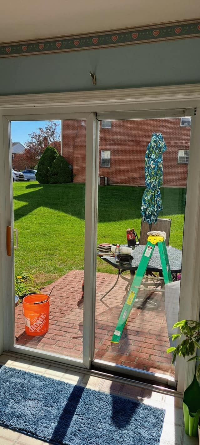 View through a sliding glass door; yard with grass and patio, construction materials, ladder, and a blue rug.