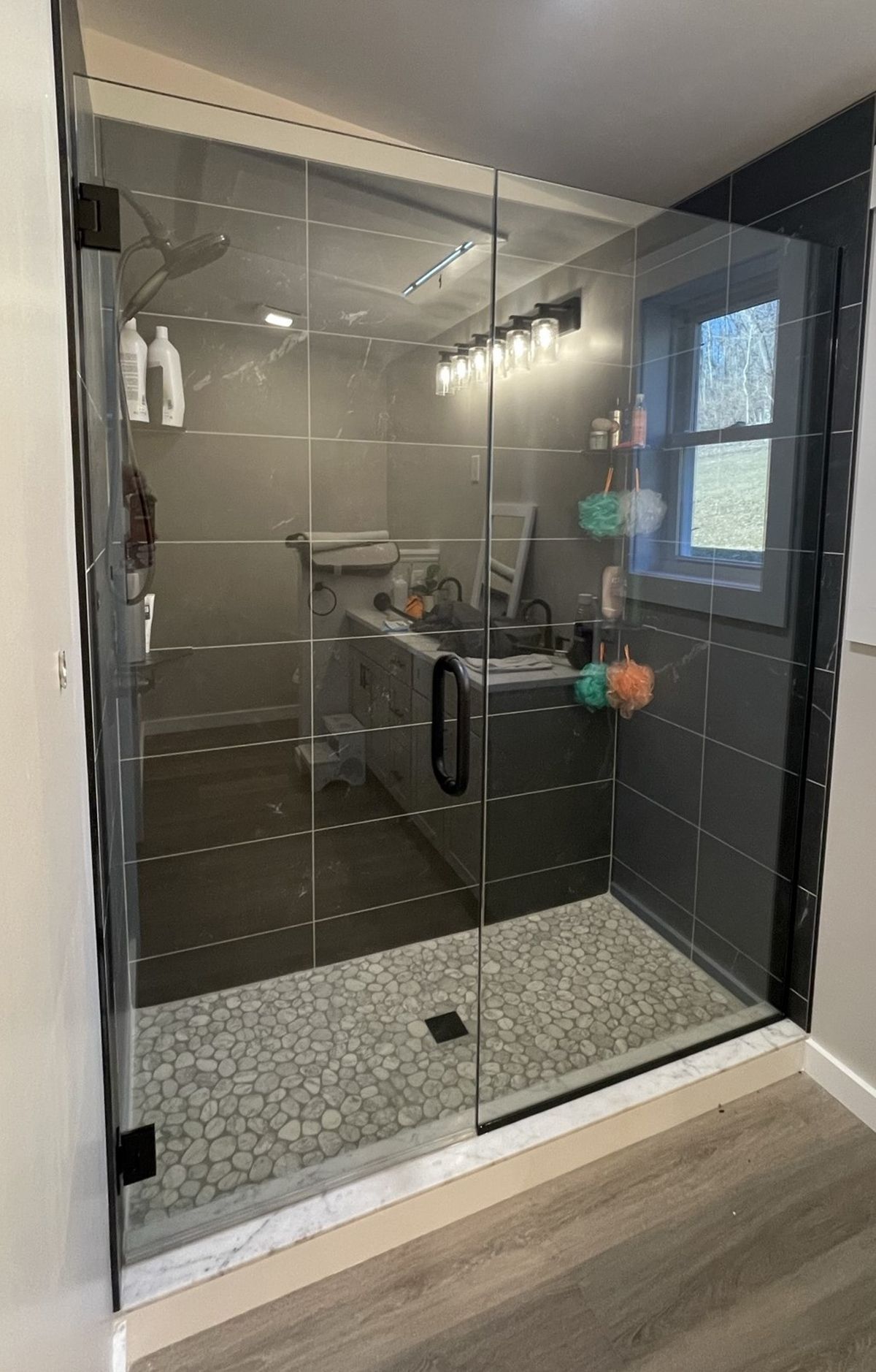 Modern glass shower with pebble floor, gray tile walls, dark hardware, and a window.