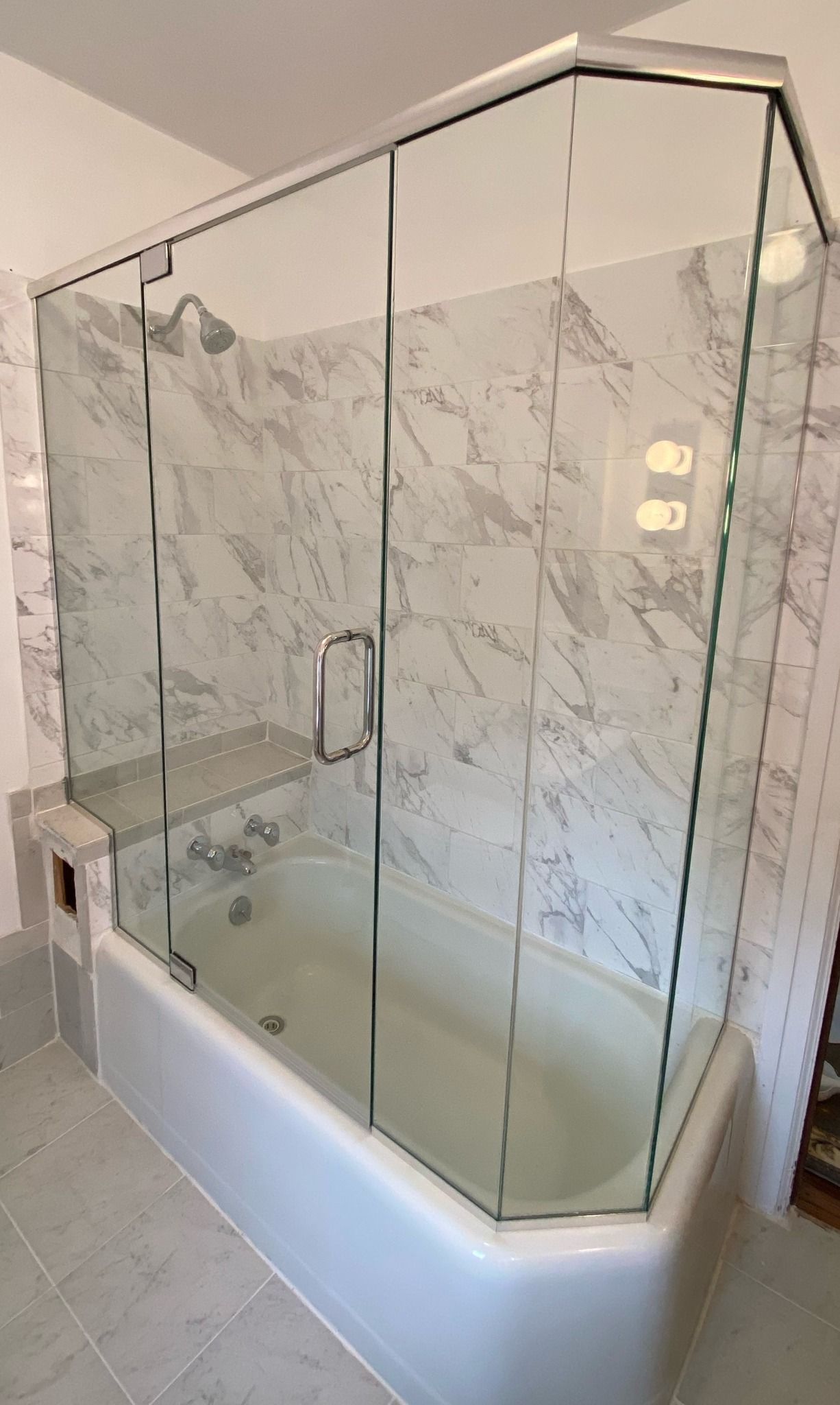 Glass shower enclosure over a white bathtub with marble-look tile walls.